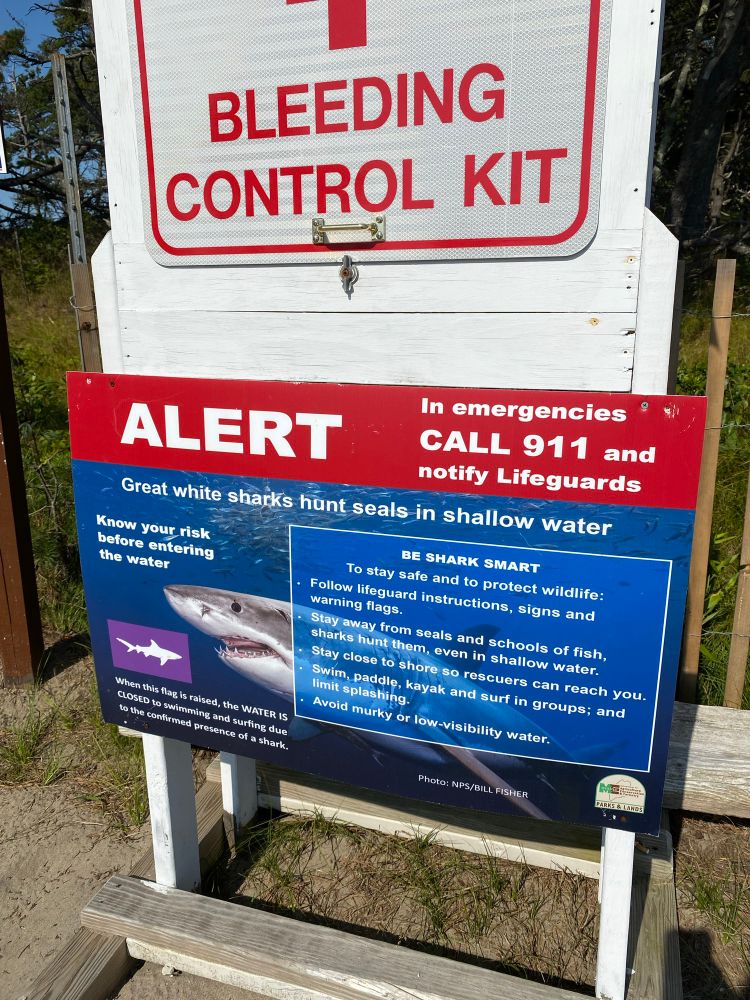 A sign near the entrance to Popham Beach in Phippsburg, ME warning of great white sharks. Above is a sign marked “bleeding control kit”.