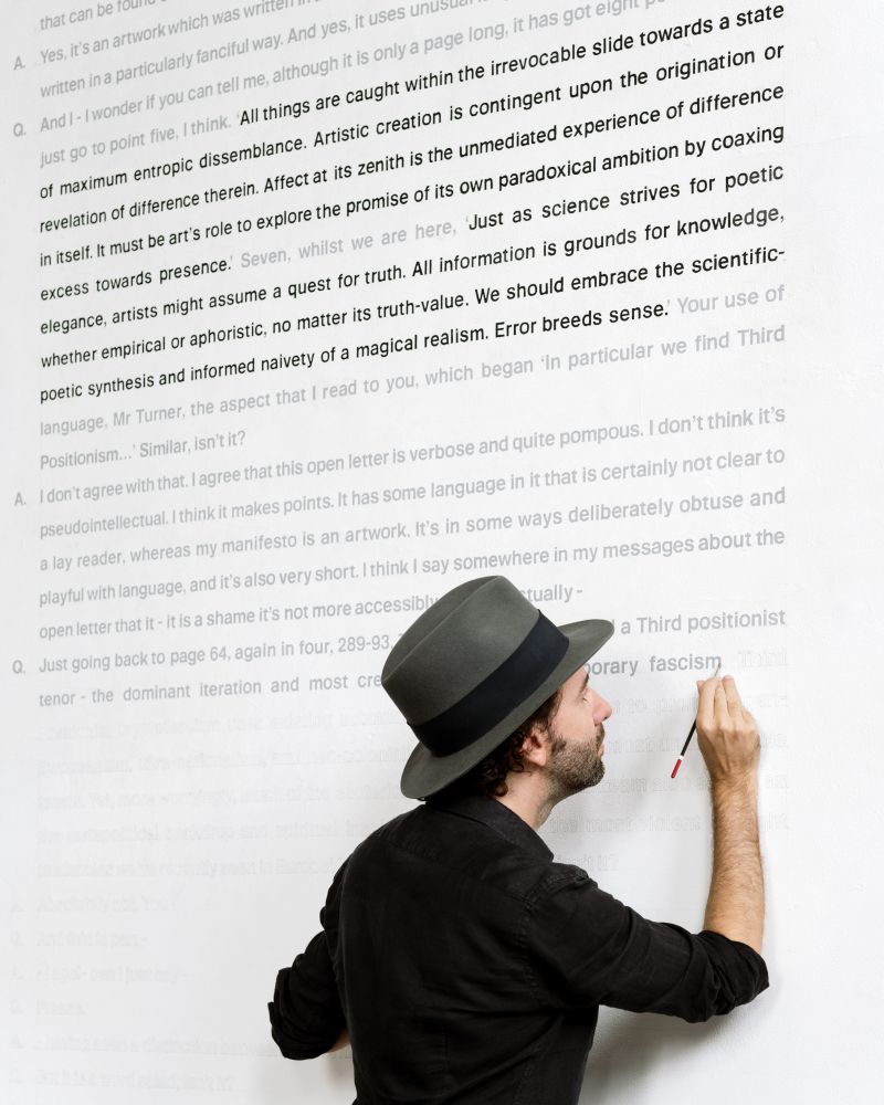 Luke painting a transcript on the gallery wall that reads:

Q. And I - I wonder if you can tell me, although it is only a page long, it has got eight points, but I will just go to point five, I think. ‘ All things are caught within the irrevocable slide towards a state of maximum entropic dissemblance. Artistic creation is contingent upon the origination or revelation of difference therein. Affect at its zenith is the unmediated experience of difference in itself. It must be art’s role to explore the promise of its own paradoxical ambition by coaxing excess towards presence.’ Seven, whilst we are here, ‘ Just as science strives for poetic elegance, artists might assume a quest for truth. All information is grounds for knowledge, whether empirical or aphoristic, no matter its truth-value. We should embrace the scientific-poetic synthesis and informed naivety of a magical realism. Error breeds sense.’ Your use of language, Mr Turner, the aspect that I read to you, which began ‘In particular we find Third Positionism…’ Similar, isn’t it?

A. I don’t agree with that. I agree that this open letter is verbose and quite pompous. I don’t think it’s pseudointellectual. I think it makes points. It has some language in it that is certainly not clear to a lay reader, whereas my manifesto is an artwork. It’s in some ways deliberately obtuse and playful with language, and it’s also very short. I think I say somewhere in my messages about the open letter that it - it is a shame it’s not more accessibly written, actually -

Q. Just going back to page 64, again in four, 289-93. This, ‘ In particular, we find a Third Positionist tenor - the dominant iteration and most creeping form of contemporary fascism…"