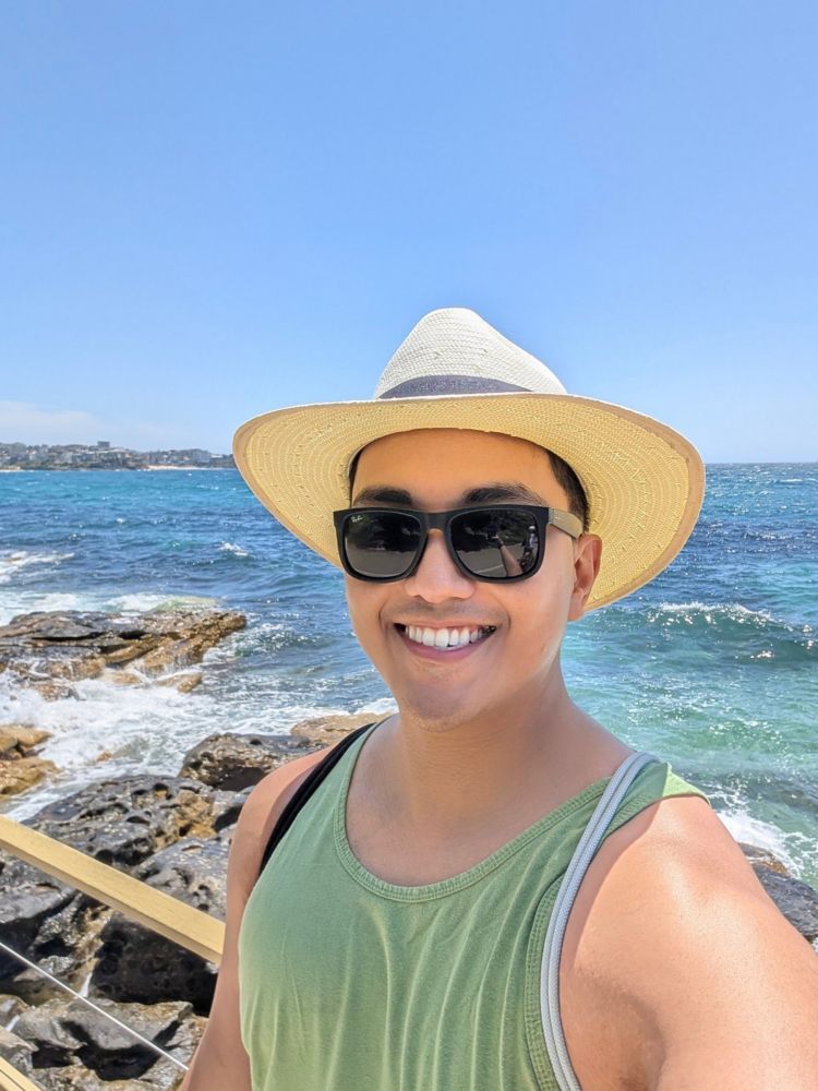 A photo of a male subject smiling, wearing a light yellow sun hat, green tank top, black sunglasses, and a drawstring bag with black strings on the right shoulder and grey strings on the left shoulder, in front of a railing by the ocean with waves crashing into rocks in the background, on a sunny, clear blue sky, kind of day.