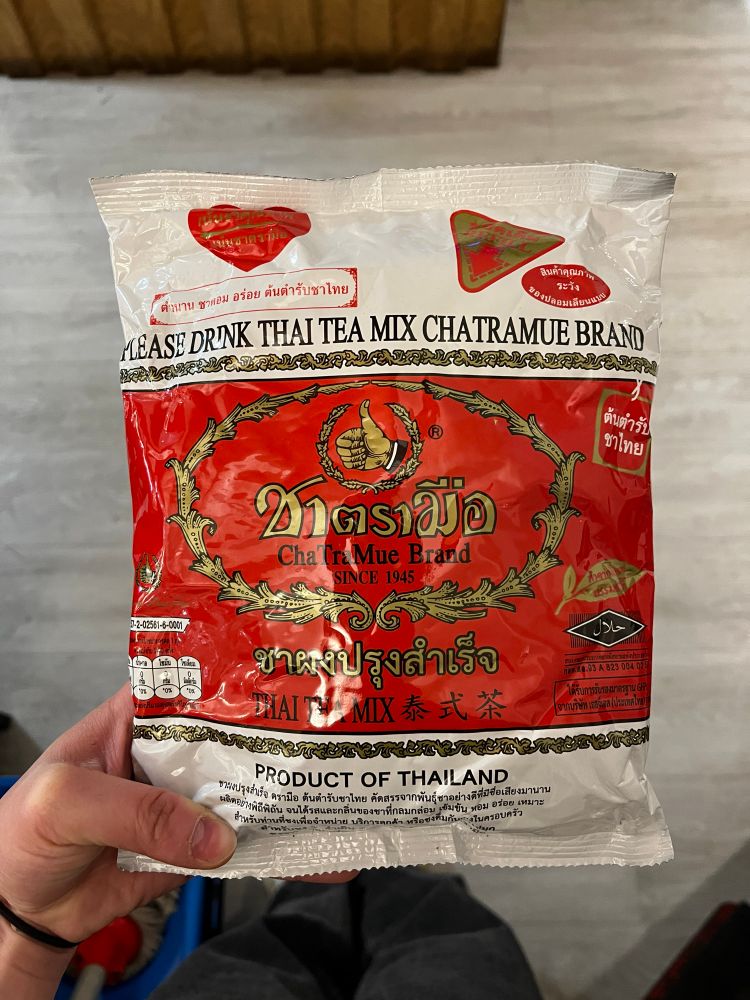 Picture of a tea bag, held in hand. It’s from ChaTraMue Brand Thai Tea, product of Thailand. White base with a red band in the centre, ornamental filigree in gold with black stroke decorates the design. Text and labels surround the front of the design.