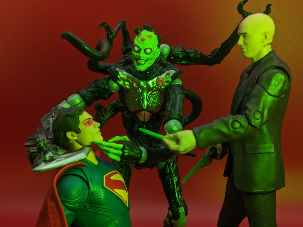 Superman is on his knees in a red room. Brainiac has a hook around his neck and is stroking his chin. Lex Luthor holds a shard of kryptonite to his face, bathing him in its green glow 