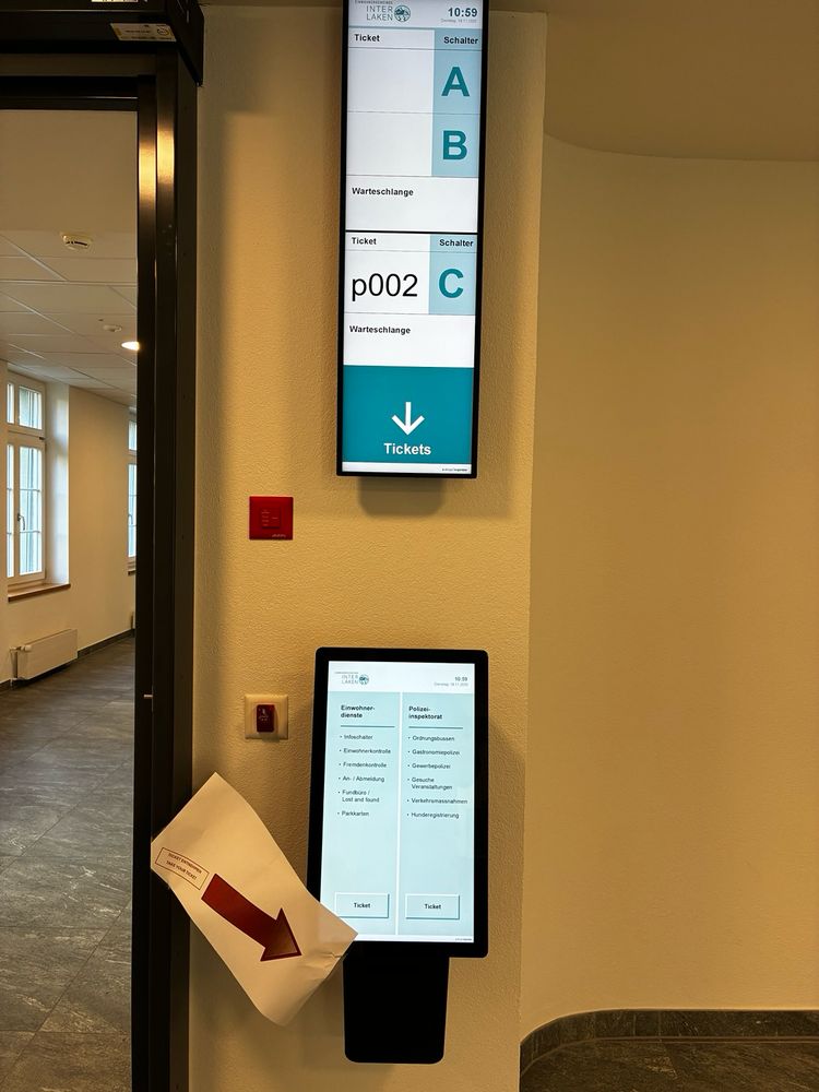 This image shows the entrance area of a municipal office, with a tall digital display mounted high on the wall and a touchscreen ticket machine below it. The upper screen shows the current time and a queue system indicating which ticket number should go to which service counter; at the moment, ticket “p002” is called to counter C. Below it, the touchscreen ticket terminal offers two columns of options: resident services such as address registration, lost and found, and parking permits on the left, and police-related services like fines, events, and dog ownership on the right. A large piece of paper with a red arrow is taped to the side of the machine, pointing to the left as an improvised sign. To the left of the ticket machine is a small red emergency button, and in the background is a hallway with windows letting in natural light.