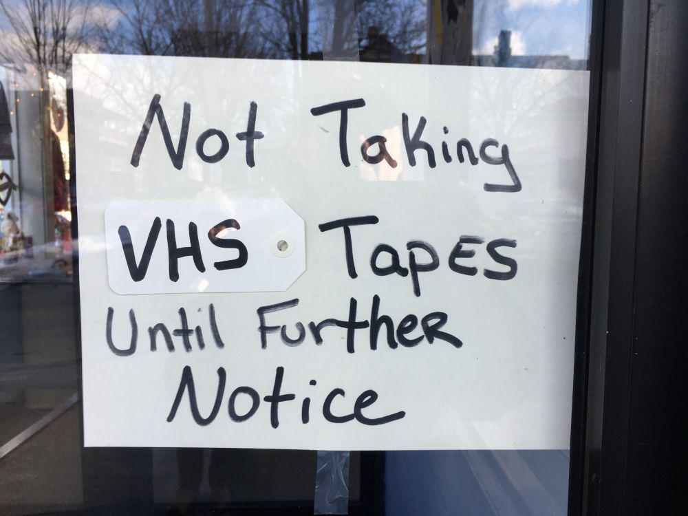 A hand-written sign in a thrift store window states that VHS tapes will not be accepted until further notice. The letters "VHS" are written on a tag that appears to be removable, suggesting that other kinds of tapes were, or could be not accepted. 