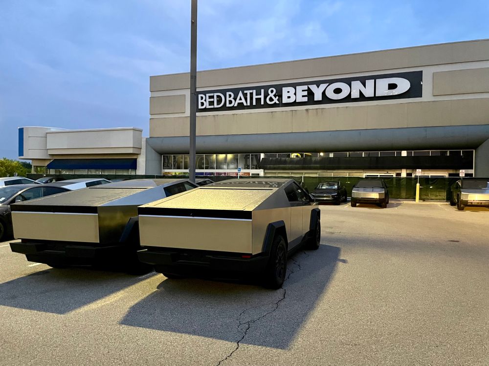 Photograph of several unsold Tesla Cybertrucks in a strip mall parking lot in the evening in front of a shuttered Bed, Bath, & Beyond store.