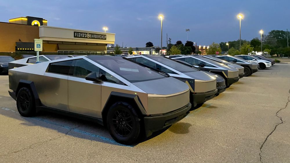 Photograph of a dozen or so unsold Tesla Cybertrucks in a strip mall parking lot in the evening. 