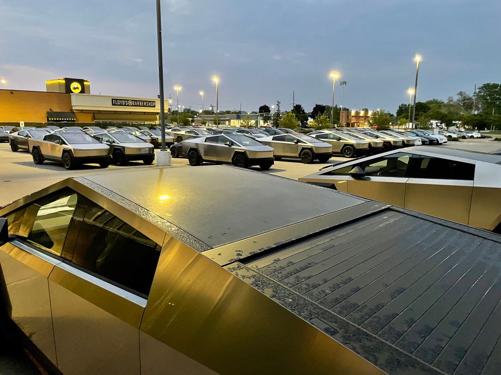Photograph of a couple dozen unsold Tesla Cybertrucks in a strip mall parking lot in the evening.