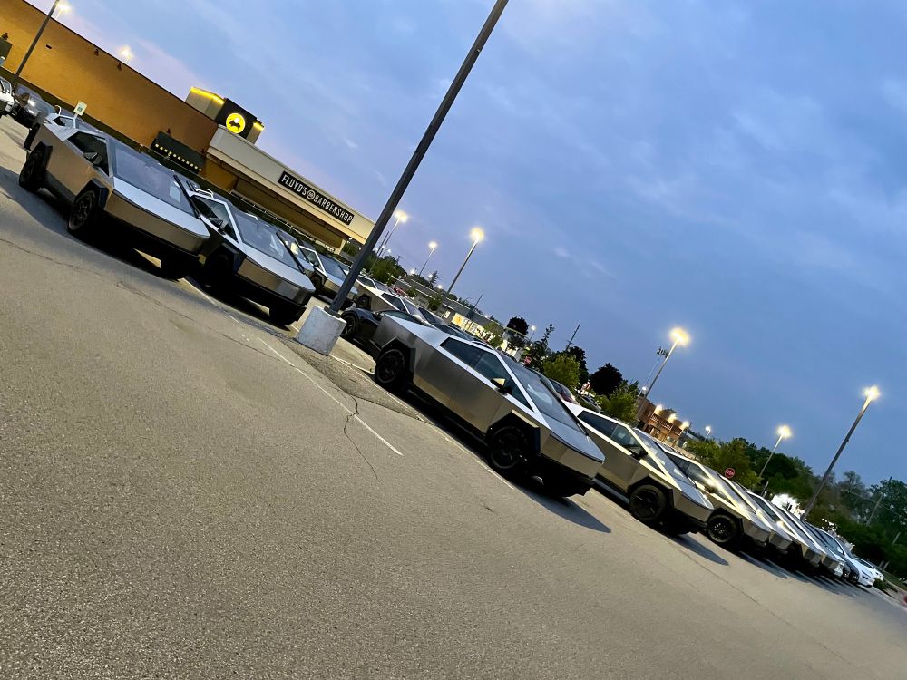 Photograph of a couple dozen unsold Tesla Cybertrucks in a strip mall parking lot in the evening. Photo slants from upper left to lower right.