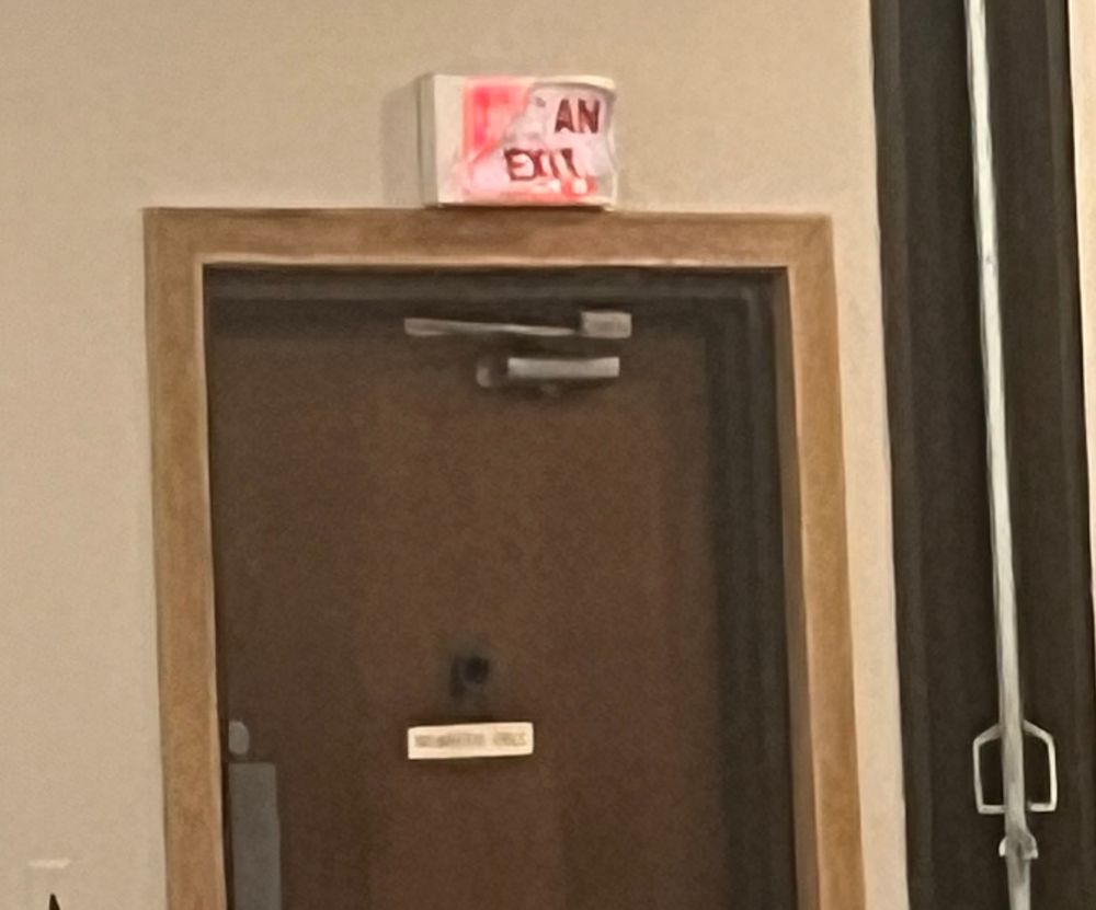 A door with an electric “EXIT” sign above, except someone taped a paper that said “NOT AN EXIT” over it, except the paper is ripped so it just reads “AN EXIT”