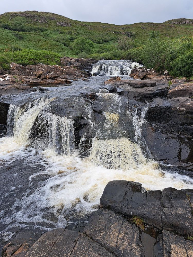 Water cascading over dark rocks with green hills in the background 