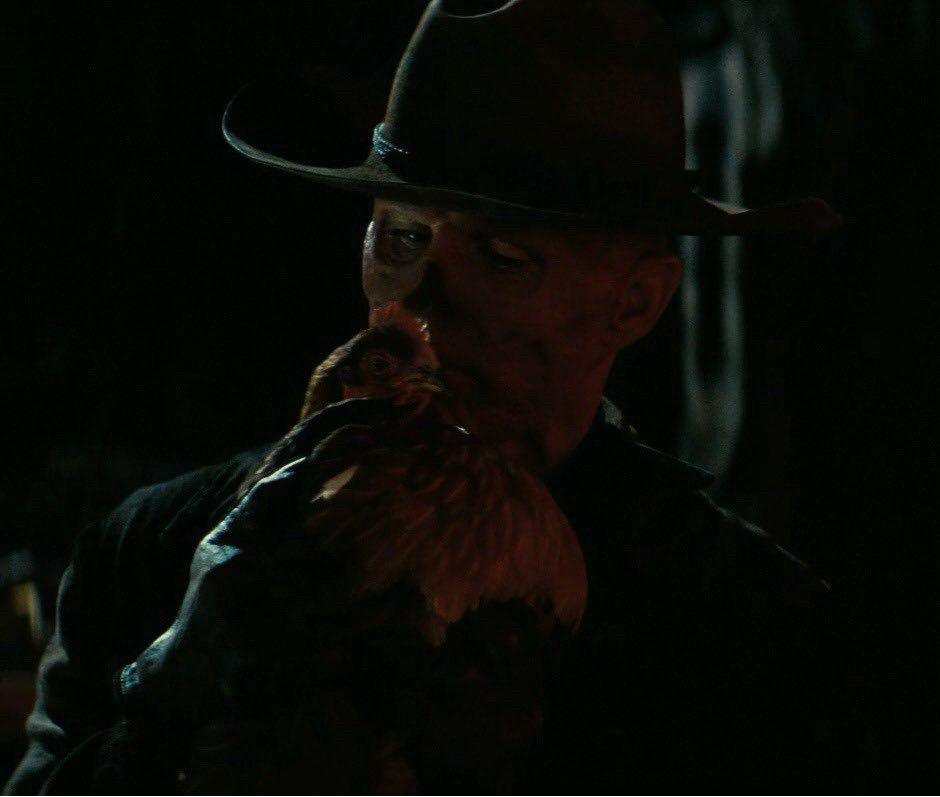 Dimly Lit Character Closeup From Fallout Series Season 1. The Ghoul Wearing a Black Cowboy Hat Whispers To A Hen