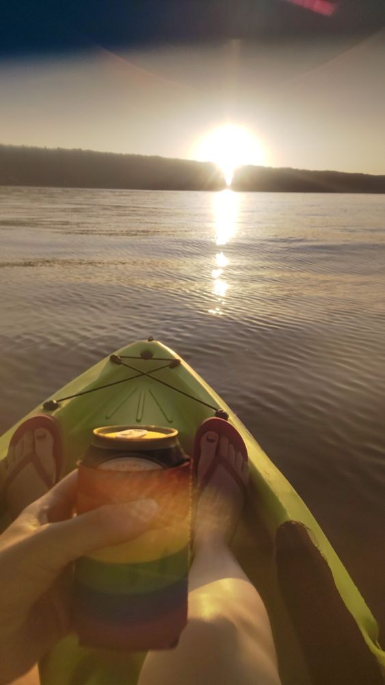 First person view from green kayak, on the water looking at the starting sunset. Person is wearing pink flip flops and holding a drink in a rainbow drink insulation sleeve.