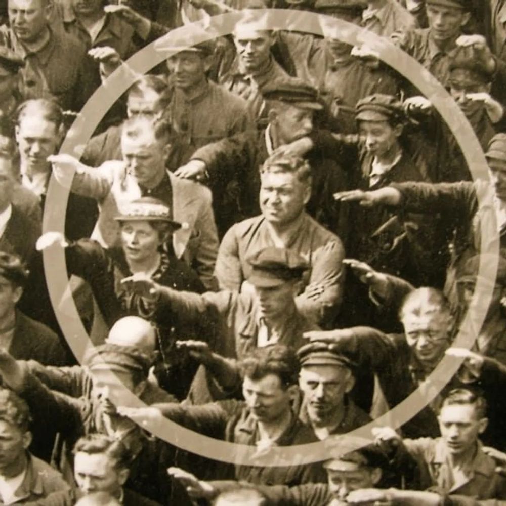 Black and white photo from 1936. August Landmesser standing with arms crossed, refusing to Nazi perform the Nazi salute in a crowd of people performing the Nazi salute. 