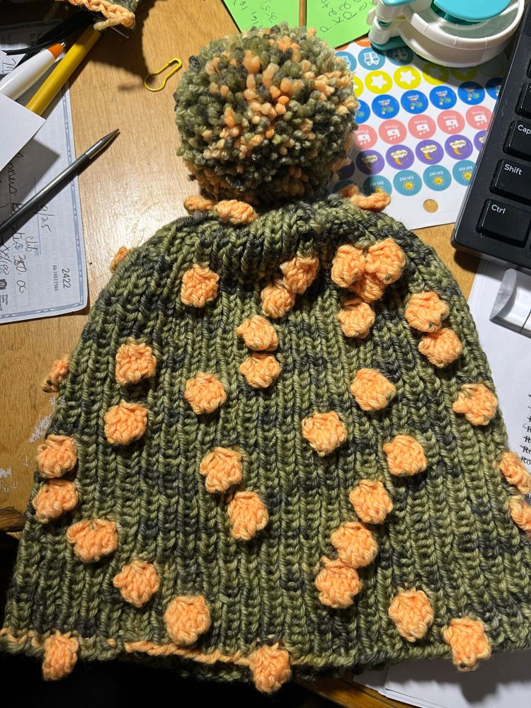 Camo beanie with yellow bobbles on a cluttered desk.