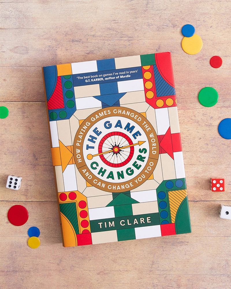 The hardback copy of The Game Changers by Tim Clare, sitting on a wooden table with colourful game pieces and dice surrounding it. The cover resembles a board game face with arrow, dots and geometric shapes in colours of blue, red, green, yellow and white.