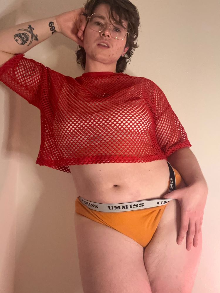 Picture of Lex, a white trans masculine person, in a red mesh top and orange thong 