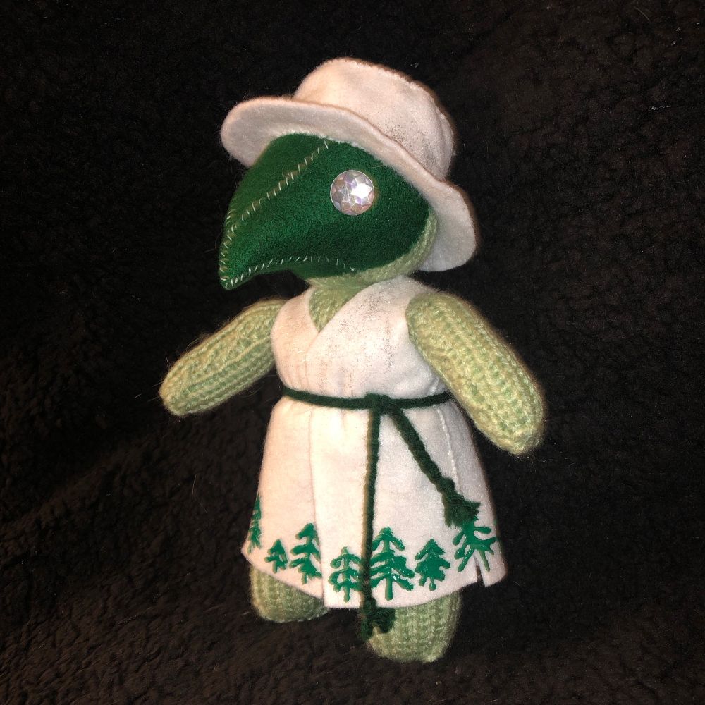 A knitted plague doctor made with light green yarn. His robes and hat are white felt and his mask is dark green felt. He has silver rhinestone eyes. A border of pine trees is painted on the hem of his robe in fabric paint. 