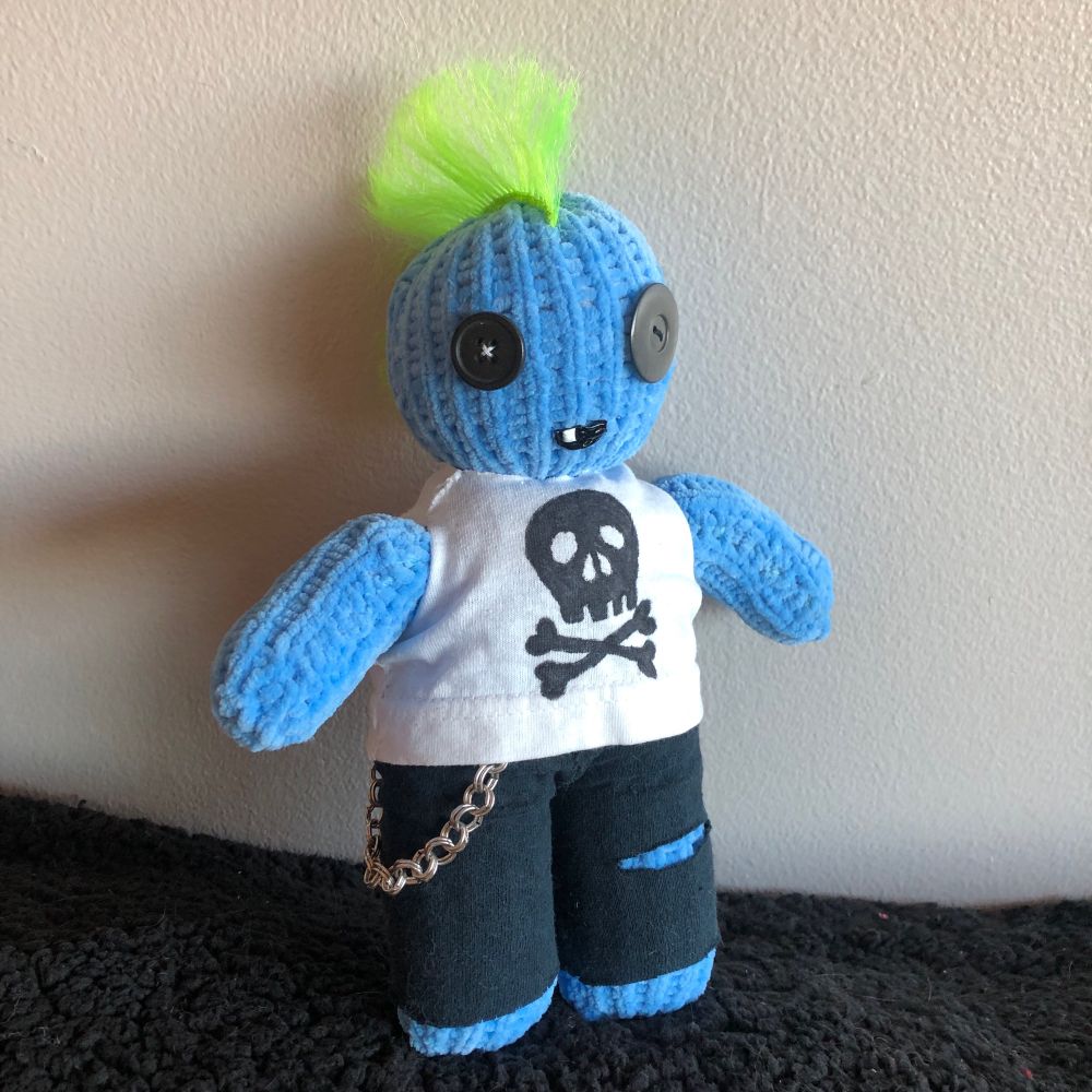A plushie zombie made with light blue yarn. He has green spiky hair and fabric clothes. Black pants, a white t-shirt with skull and crossbones, and a metal chain hanging from his pants. 