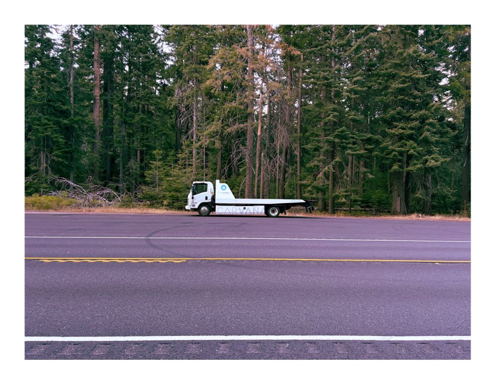 Photo of a carvana truck on a scenic road through the forest. 