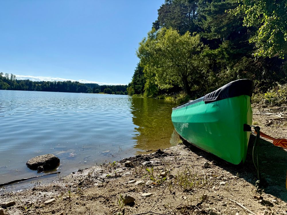 Ein steiniger Seestrand mit einem angebundenen grünen Kajak rechts.
Daneben Blick auf den See mit Bäumen im Hintergrund.