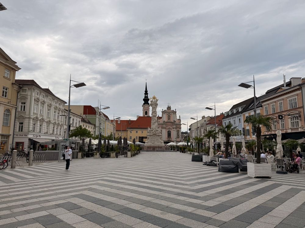 Rathausplatz in St.Pölten. Der Boden besteht aus hellen und dunklen Platten im Zickzackmuster. Links im Bild ist eine Frau mit Baby im Arm. In der Mitte ist bis zur Pestsäule eine große freie Fläche, darum sind Palmen in eckigen Trögen und Schanigärten. 
Im Hintergrund rechts und links sind Wohnhäuser, ganz hinten in der Mitte die Kirche. 