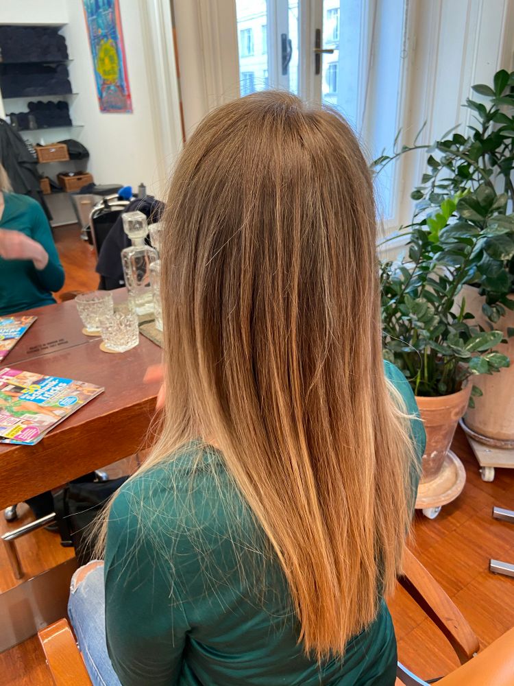 Foto vom Hinterkopf einer sitzenden Frau in grünem Shirt mit langen Mittelblonden Haaren bis Mitte des Rückens.
Im Hintergrund ist Links ein Spiegel, rechts zwei Pflanzentöpfe auf Holzboden. 