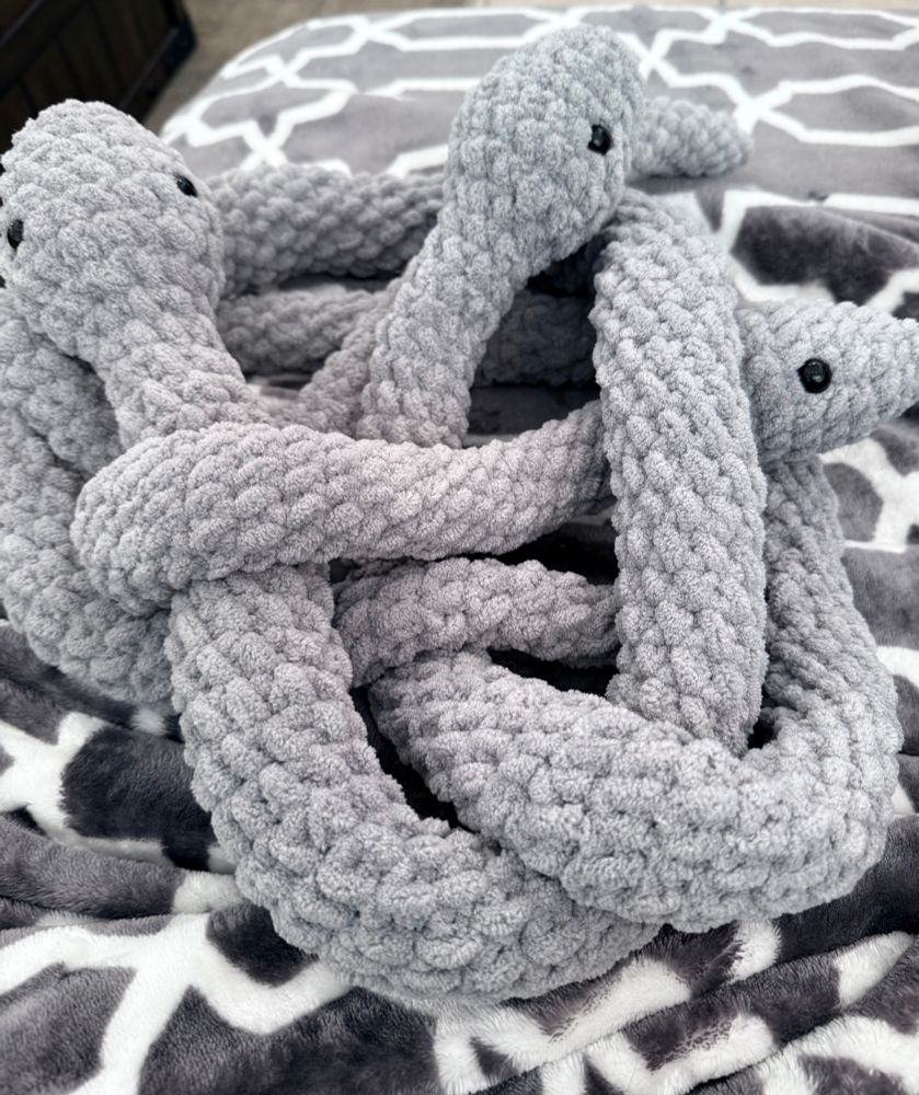Three gray crochet snakes with black eyes twisted together in a pile on a gray and white blanket.