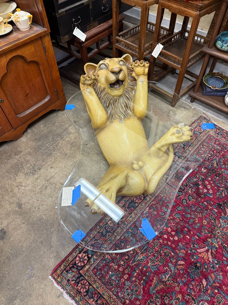 Glass table with wonderfully goofy yellow lion holding it up with entire belly exposed in antique shop