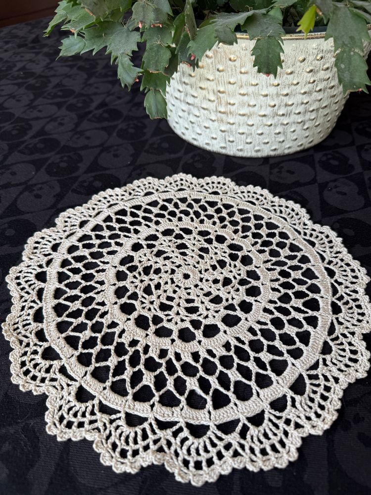 Ecru round crochet lace doily on black table cloth next to a Christmas cactus plant in a white pot.