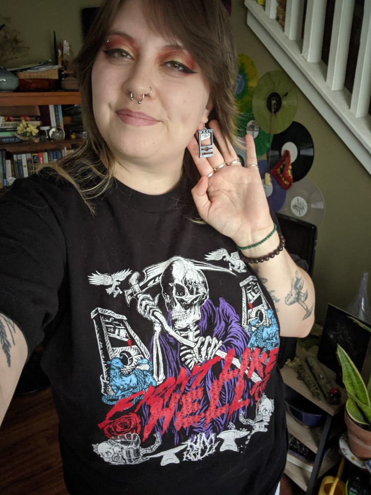 me, a white femme, wearing my Fight Like Hell t-shirt that features a skeleton with a scythe and some guillotines, plus some eye makeup and guillotine earrings