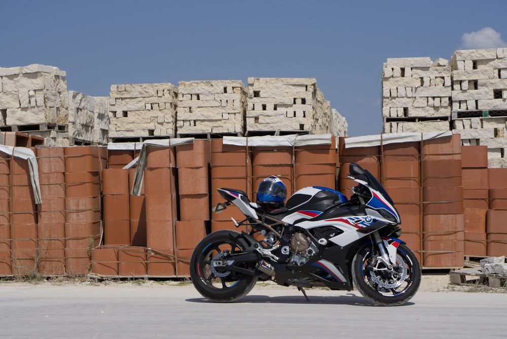 A photo of a white, blue, and red painted BMW S1000RR sport bike on its kickstand in a brick-yard. There are stacks of masonry bricks and pavers occupying the entire horizon.