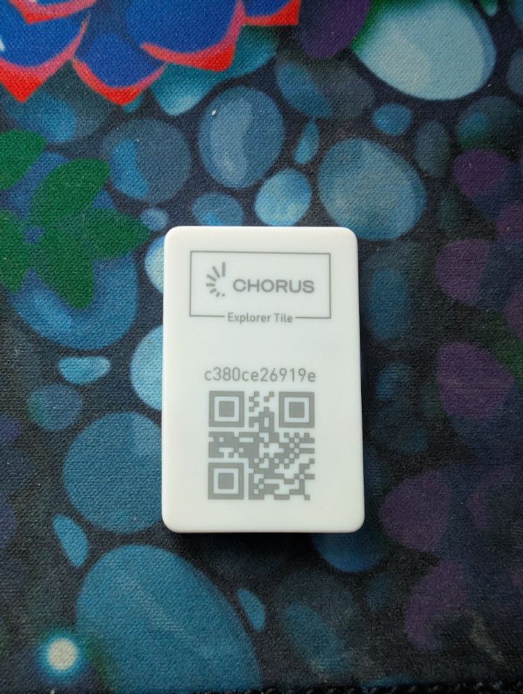 a photo of a small while rectangular device not much larger than a quarter in size sitting on a desk. the device has some printing on the front that says CHORUS Explorer Tile and 'c380ce26919e' along with a QR code sharing the same string