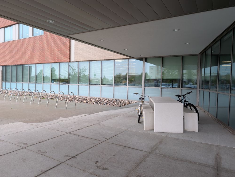 Two bicycles locked to a picnic table under the shelter of a cantilevered roof, while the bike rack is just to the left, unsheltered in the rain