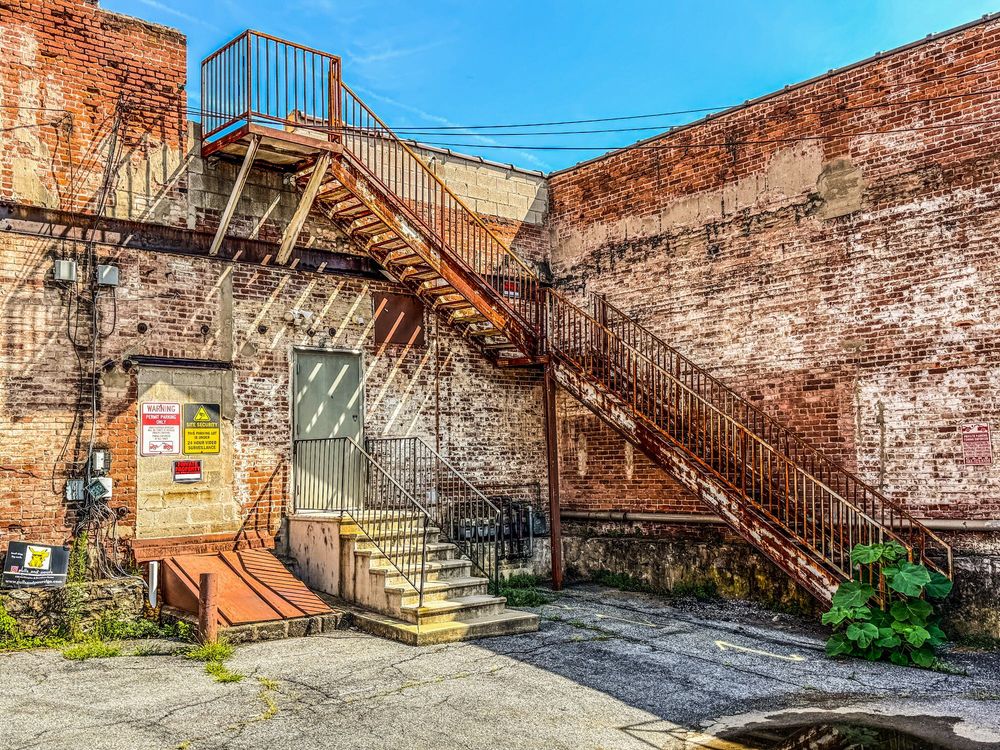 A rooftop fire escape on a brick building goes down one flight to an adjacent brick wall and turns 90° then down to ground level on the adjacent wall. Below the fire escape there is a door and stairway going into a parking lot. To the left of the door there is a cellar entrance with security and warning signs mounted on the wall above.