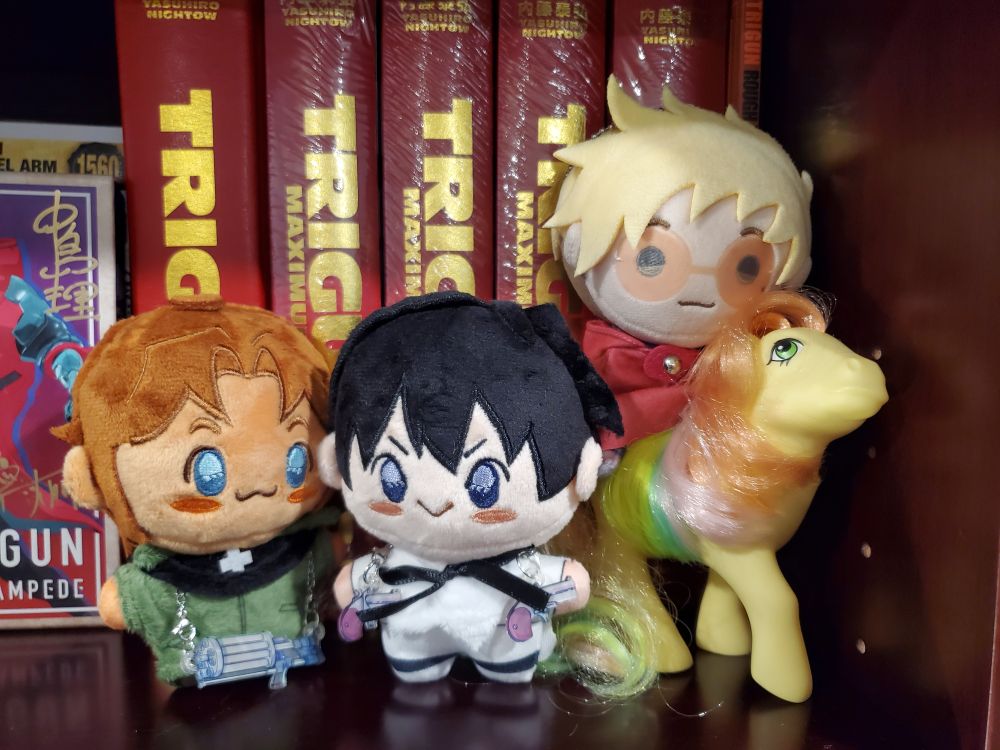 A photo of three plushies, two depicting the Trigun manga versions of Milly Thompson (left) and Meryl Stryfe (middle) with their weapons, the third plushie (right) is a Teru Vash (aka Teruva) plushie depicting the Trigun Stampede version of Vash the Stampede. Teru Vash is sitting in top of a yellow G1 (generation 1) My Little Pony from the 1980s. Behind them are the Trigun and Trigun Maximum deluxe edition mangas published by Dark Horse and an autographed Trigun Stampede Blu-ray collection. 