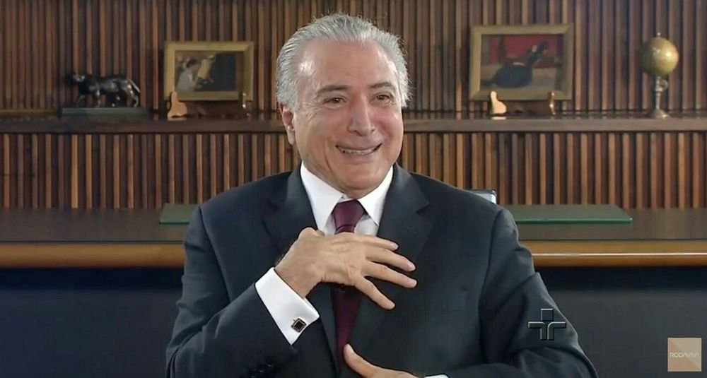 foto do ex-presidente michel temer, um homem branco de cabelos idem, usando terno escuro, sorrindo e com a mãozinha espalmada sobre o próprio peito 