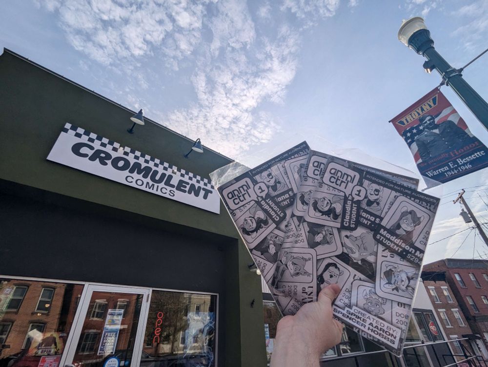 A sunny blue sky with wispy clouds, above a street with stores and brick buildings. The focus is on Cromulent Comics. There is a lightpole with a flag that says TROY NY. A white man's hand holds up three copies of issue 1 of Anml Ctrl
