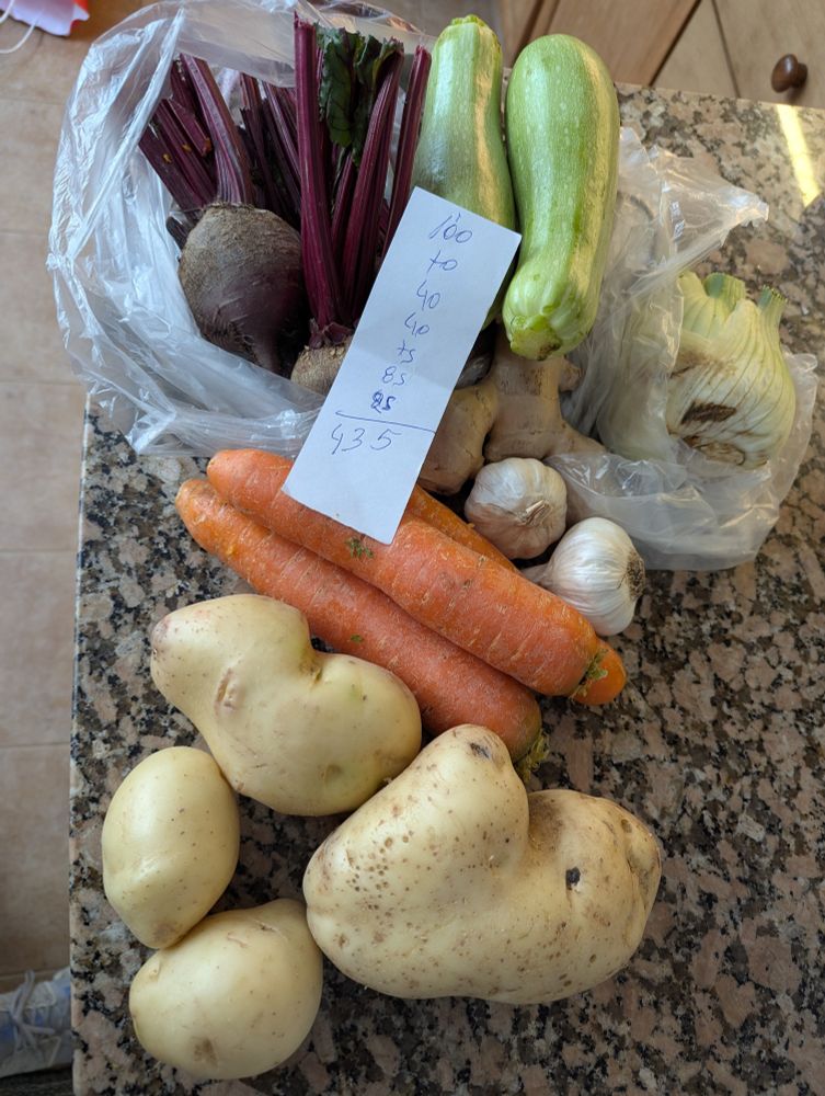 Beets, fennel, carrots, postales, garlic, ginger on a counter with a piece of paper that has numbers that add up to 435 (UY pesos)