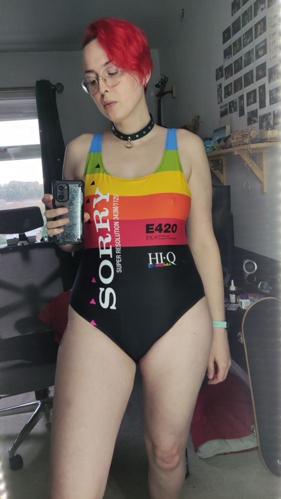The pale fem Rach queer with vivid red short hair wearing their round glasses and a dark green collar with silver studs and a silver ring on it and a colourful rainbow swimsuit that directly evokes the style of OG VHS tape box designs and it says SORRY, E420 HI-Q