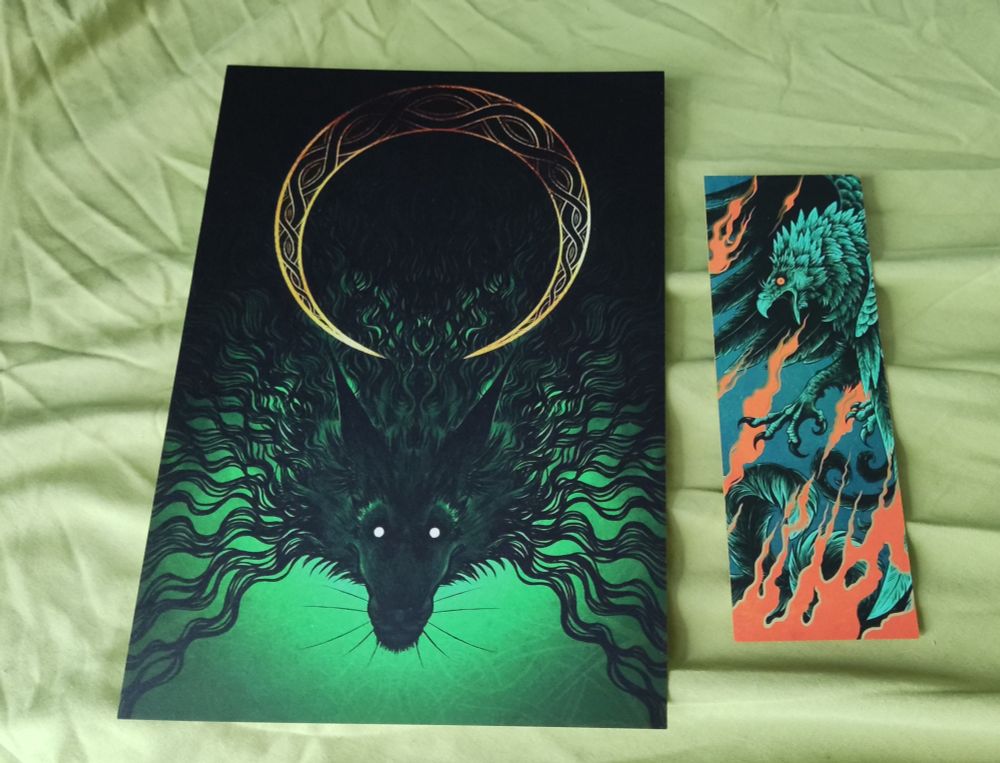 An a5 print of a dark black wolf design on dark green with a golden circle, and a bookmark of a bird shooting fire