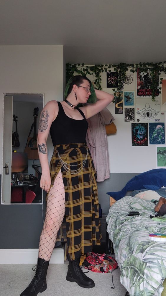 Rach! A pale fem queer with vivid green hair standing to one side whilst wearing black boots, fishnets, a cute black and yellow skirt, black tank top black choker, and some silver accessories