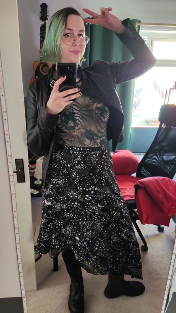 Rach! A pale fem queer with vivid green hair standing whilst wearing a green lacy bodysuit, black starry skirt, black boots, and a black leather jacket