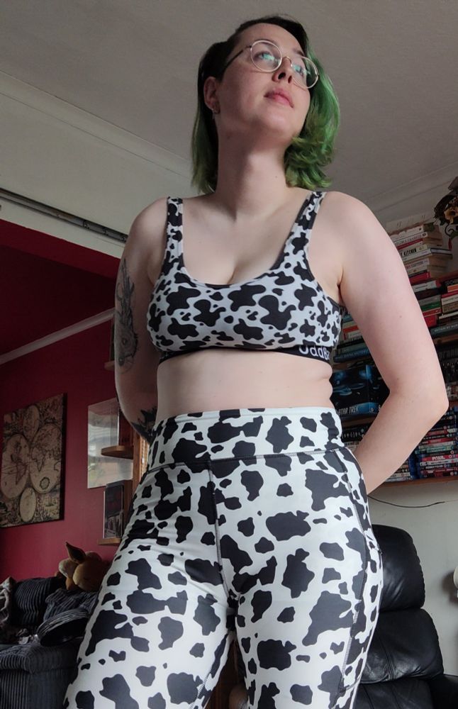 Rach! A pale fem queer with vivid green hair standing whilst wearing cowprint bra and leggings