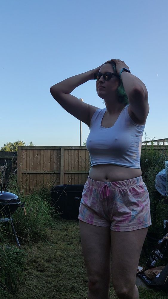 Rach! A pale fem queer with vivid green hair standing in a garden wearing soft pastel shorts and a white crop top straining to contain huge tits