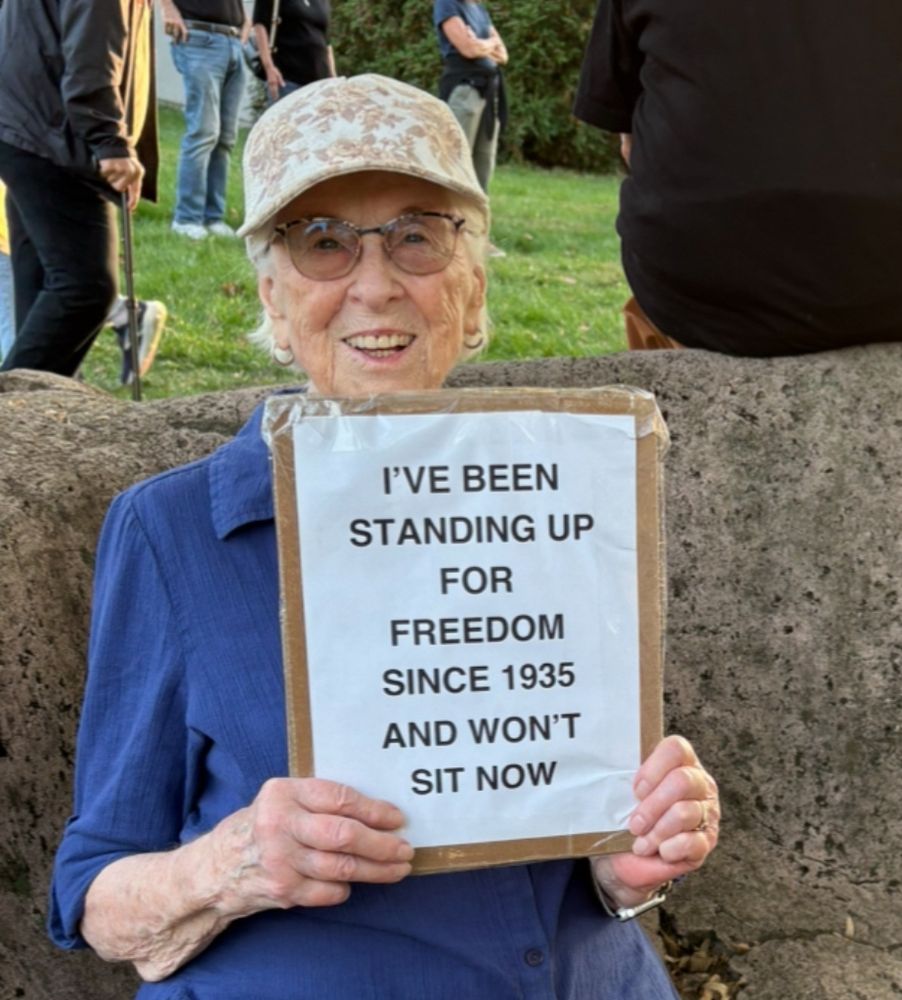 Elderly woman in a hat standing outside with a sign that says 

I'VE BEEN
STANDING UP FOR
FREEDOM
SINCE 1935
AND WON'T
SIT NOW