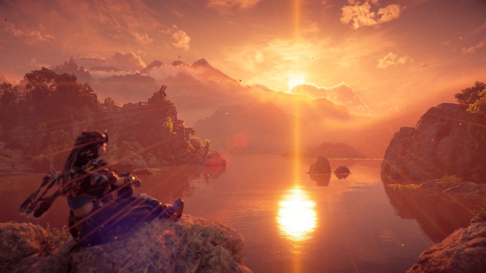 A beautiful sunset peaks over the mountains and reflects in a large river. Aloy is on the left side sitting down and enjoying the view.