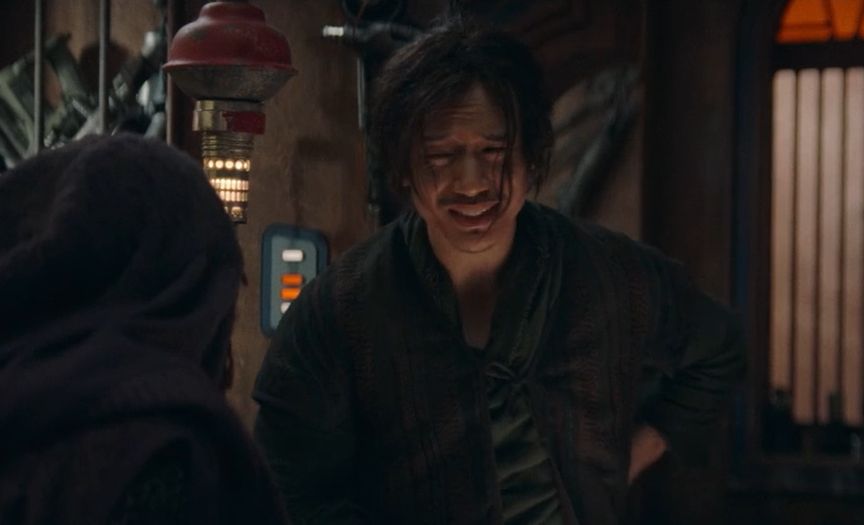 qimir in the apothecary talking to mae about her jedi killing plans with a scrunched up, judgemental face, hand on hip and hair in face.