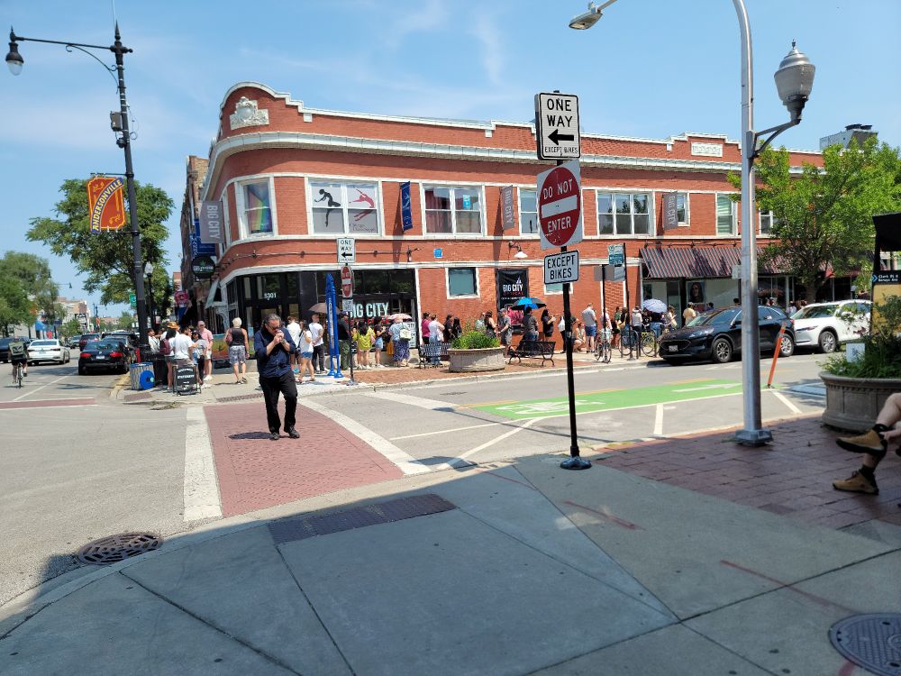 A street corner with a line of people wrapping around the corner on a sunny Saturday morning.