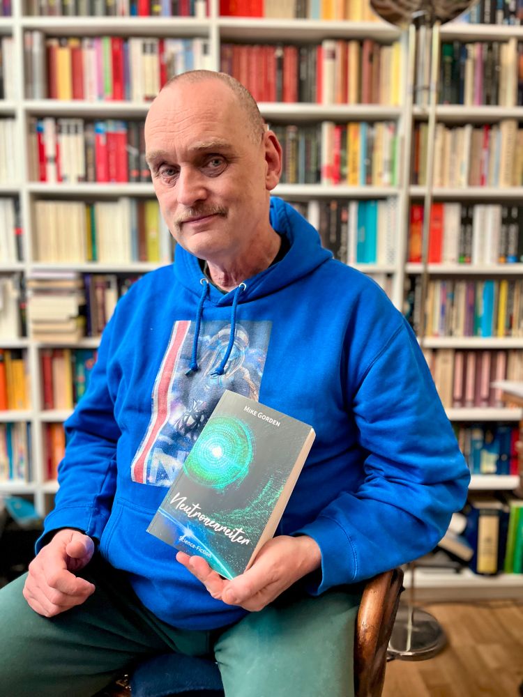 Photo des deutschen Science-Fiction Schriftstellers Mike Gorden vor einer Bücherwand sitzend. In der Hand hält er sein aktuelles Buch »Neutronenreiter«.