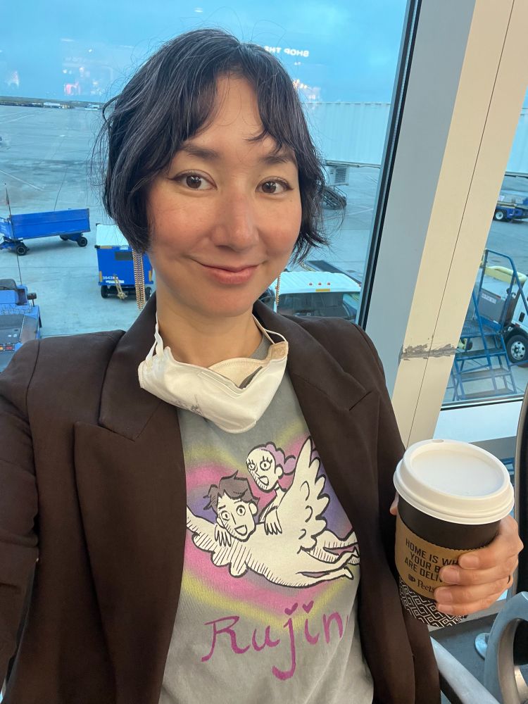 Me in an airport with a coffee, wearing a Rujinu shirt from KPop demon hunters