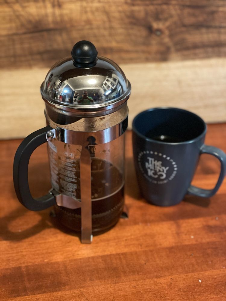 French Press Carafe-partially filled with coffee. Next to a steaming mug filled with black coffee. 